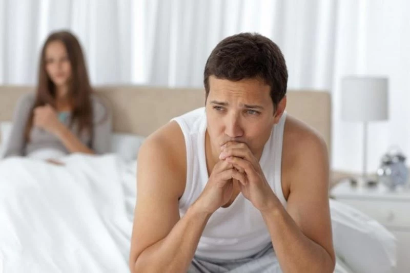 Chồng ngồi buồn bã ngồi cạnh giường, phía sau là người vợ thờ ơ cầm điện thoại